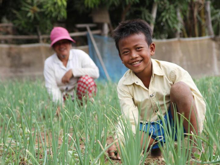 Khmer boy in the foreground kneeling in a grassy field with his mum in the background