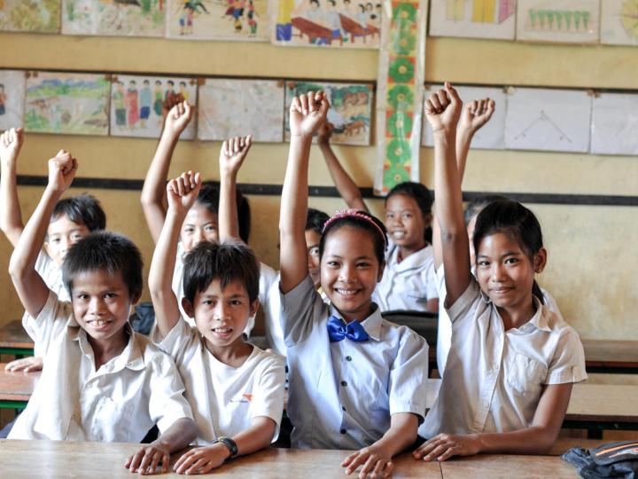 Group of Khmer students smiling with their hands raised in the air sitting at desks inside a classroom